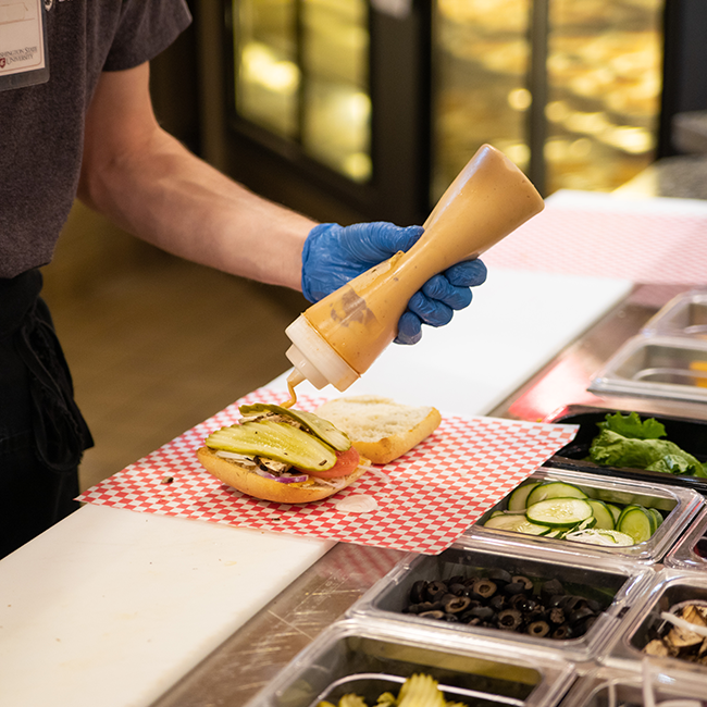 An employee putting mustard on a sandwich. 